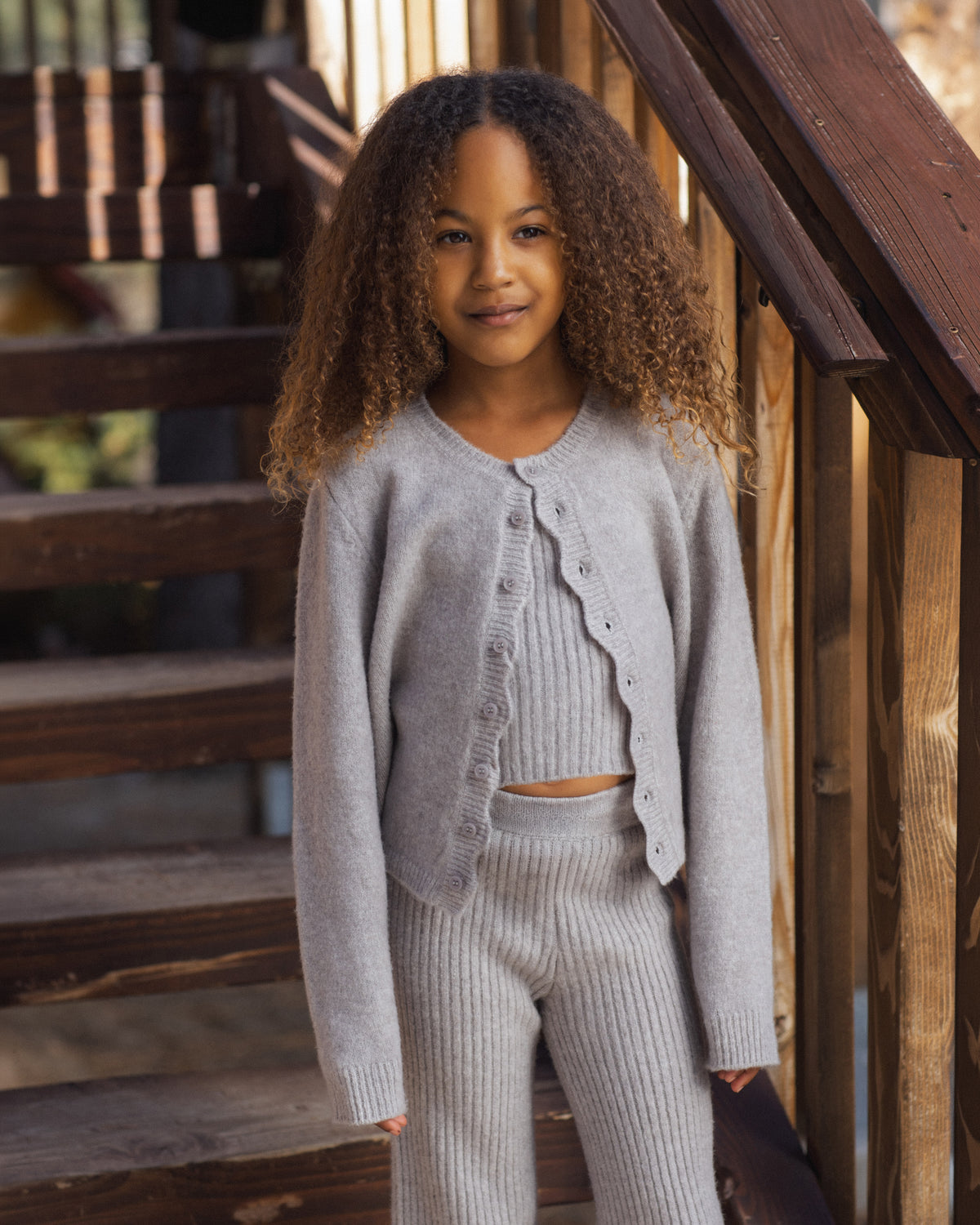 Child wearing a grey knitted sweater and matching pants and top standing in front of an exterior wooden staircase.