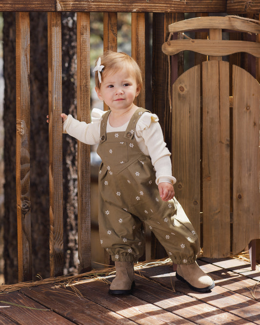 A toddler girl wearing a brown floral overalls outfit over a white long-sleeve shirt, standing on a wooden deck in front of a wooden fence.
