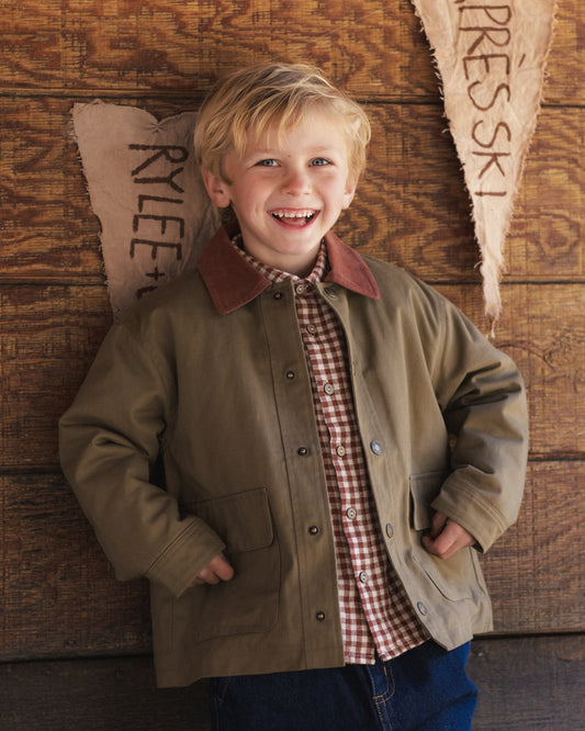 Child wearing a olive coat with a brown collar leaning against a wooden wall