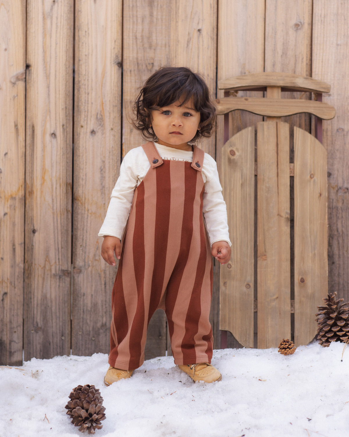 A toddler stands in snow in front of a wooden fence, wearing rust and tan vertically striped overalls over a cream long-sleeve shirt and tan boots.