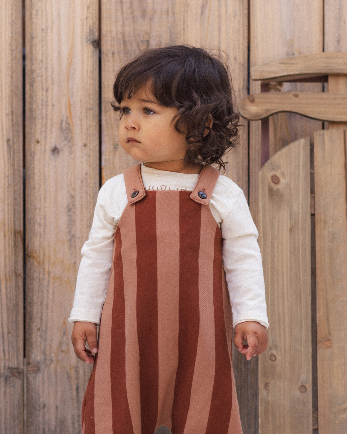 A toddler with dark curly hair wearing a white long-sleeve shirt and a rust-brown and dusty pink vertically striped pinafore overalls, standing in front of a rustic wooden fence.
