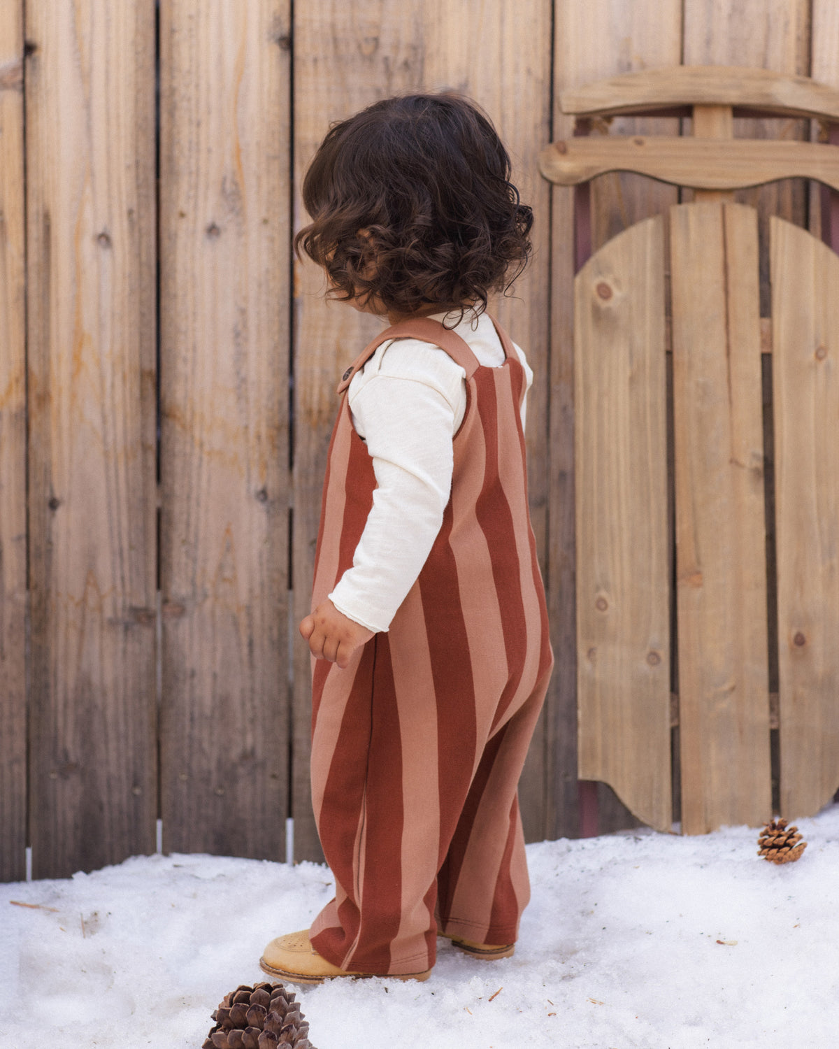 A toddler standing outdoors in front of a wooden fence and snow, wearing a rust and dusty pink vertically striped overall jumpsuit over a white long-sleeve shirt.