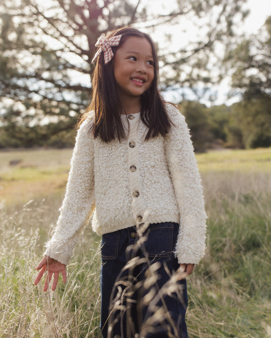 A young girl wearing a cream-colored textured knit cardigan and dark patterned pants, standing in a grassy outdoor field.