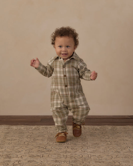 A toddler with curly brown hair wearing a beige and brown plaid button-up romper and brown leather moccasins, walking forward on a patterned rug against a beige studio backdrop.