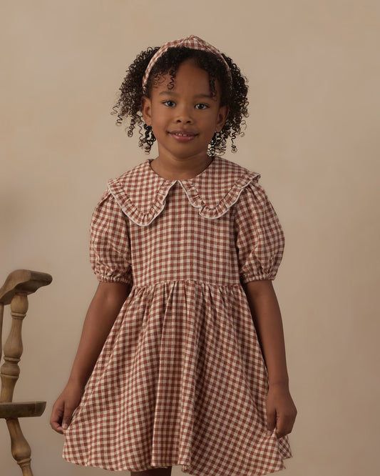 A young girl wearing a brown and beige gingham dress with a Peter Pan collar and puff sleeves, standing against a plain beige background.
