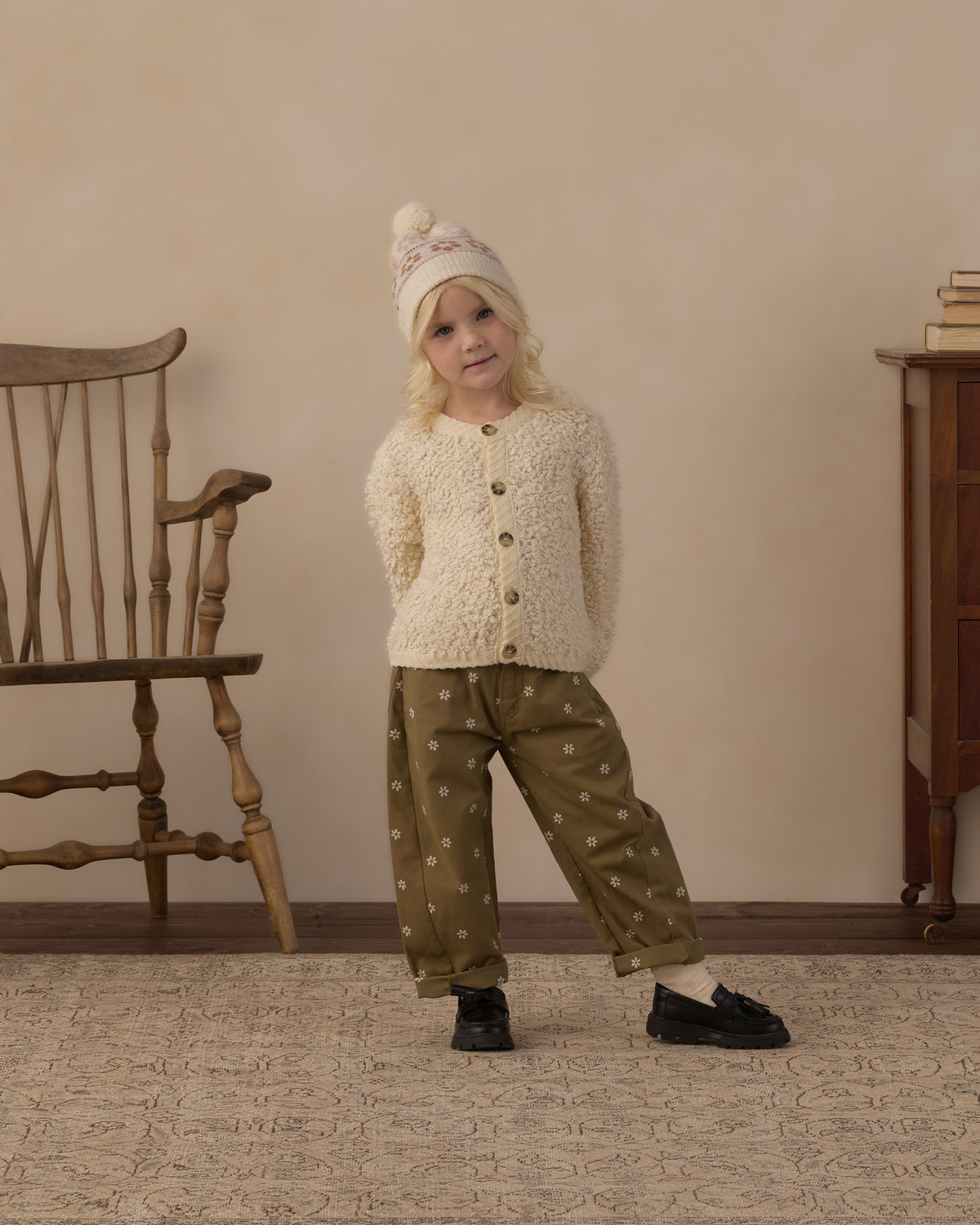 A young child stands in a room wearing a cream textured cardigan, olive green pants with a small white pattern, a cream knit hat with a pom-pom, and black shoes. The background features a beige wall, a wooden chair on the left, and a wooden cabinet on the right.