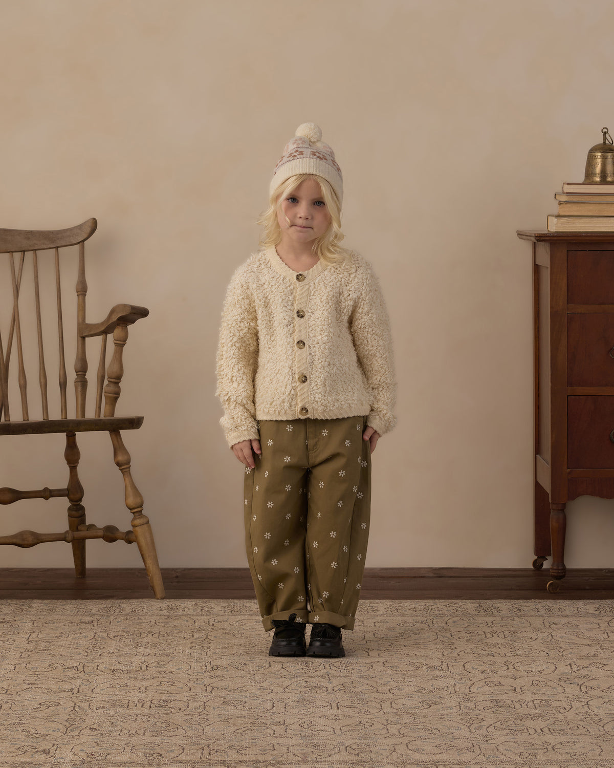 A young child stands in a room wearing a cream textured cardigan with buttons, olive green pants with white polka dots, and a cream knit beanie. A wooden chair is on the left and a wooden cabinet is on the right against a beige wall.