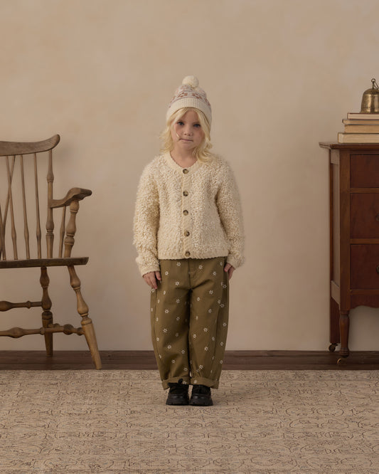 A young child stands in a room wearing a cream textured cardigan with buttons, olive green pants with white polka dots, and a cream knit beanie. A wooden chair is on the left and a wooden cabinet is on the right against a beige wall.