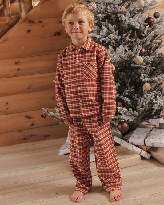 Child wearing ruby plaid two piece collared pajama set with buttons down front and right front pocket, shown in a living room with a Christmas tree