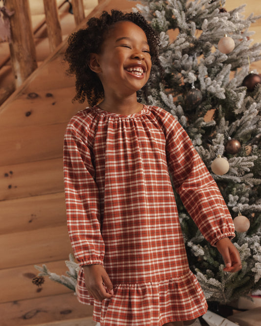 Child smiling wearing a ruby plaid nightgown with a ruffle hem standing near a Christmas tree
