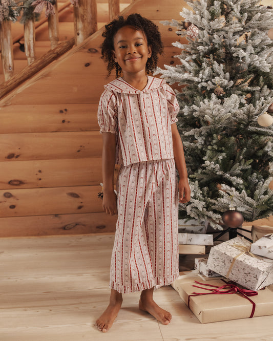 Child wearing red and white stiped two piece pajamas with a ruffle peter pan collar and ruffle detail at hem, standing in living room decorated for Christmas