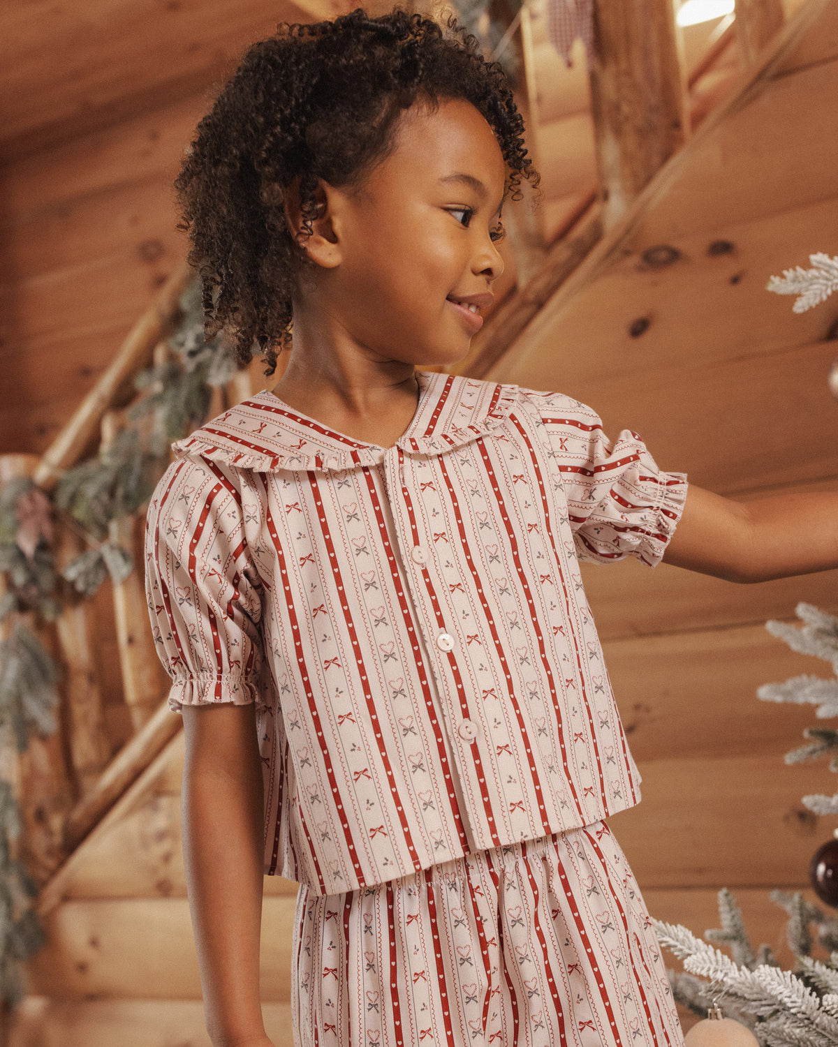 Close up of child wearing red and white stiped two piece pajamas with a ruffle peter pan collar and ruffle detail at hem, standing in living room decorated for Christmas