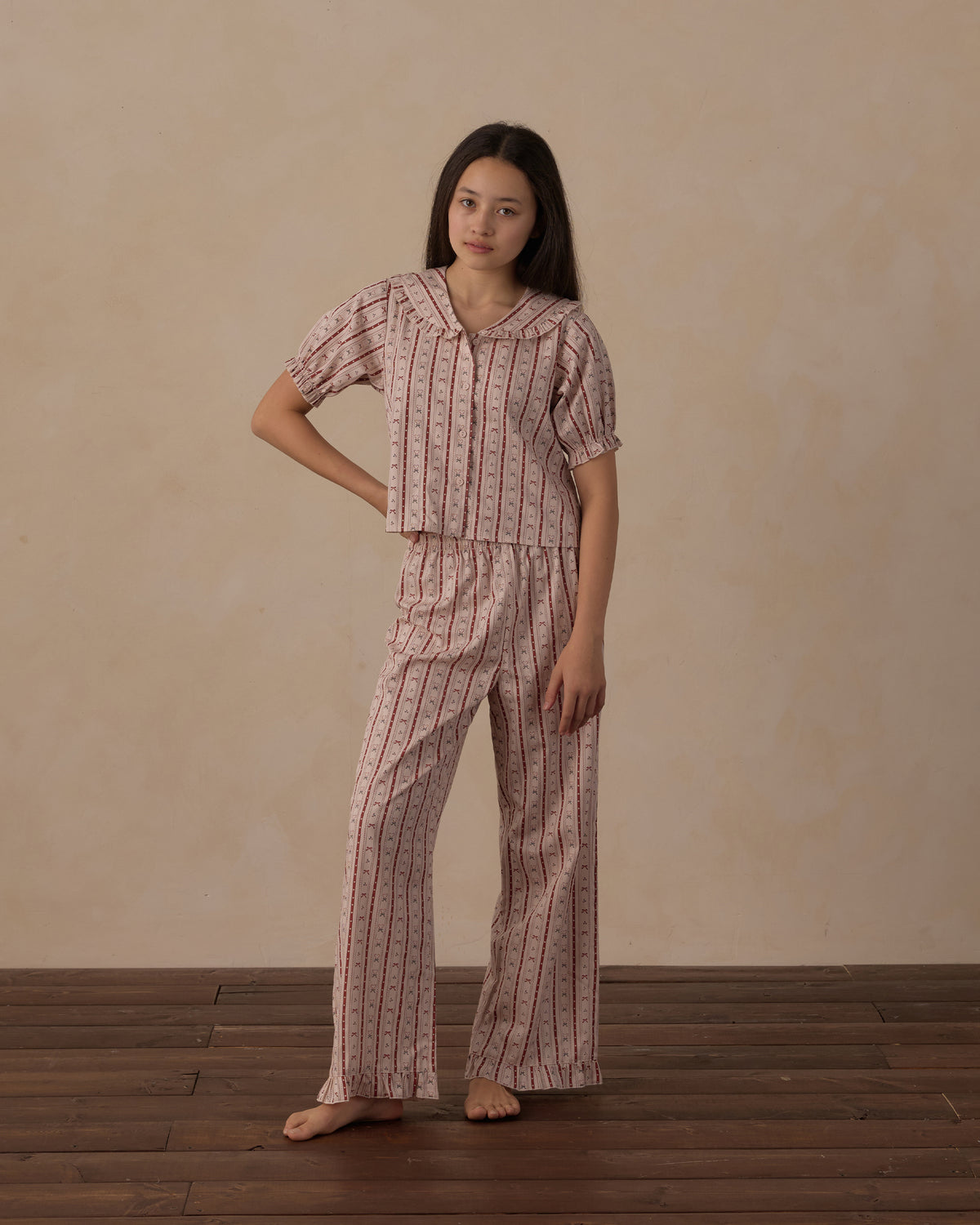 A woman stands barefoot in a studio wearing a matching short-sleeve button-front pajama top and wide-leg pants set in a light pink base with thin vertical dark red stripes and a small floral print.