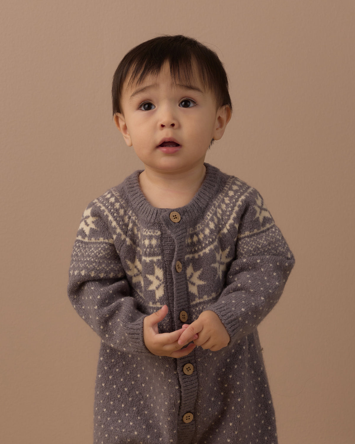 Baby wearing a grey fair isle knit romper with buttons down the front, in front of a beige background