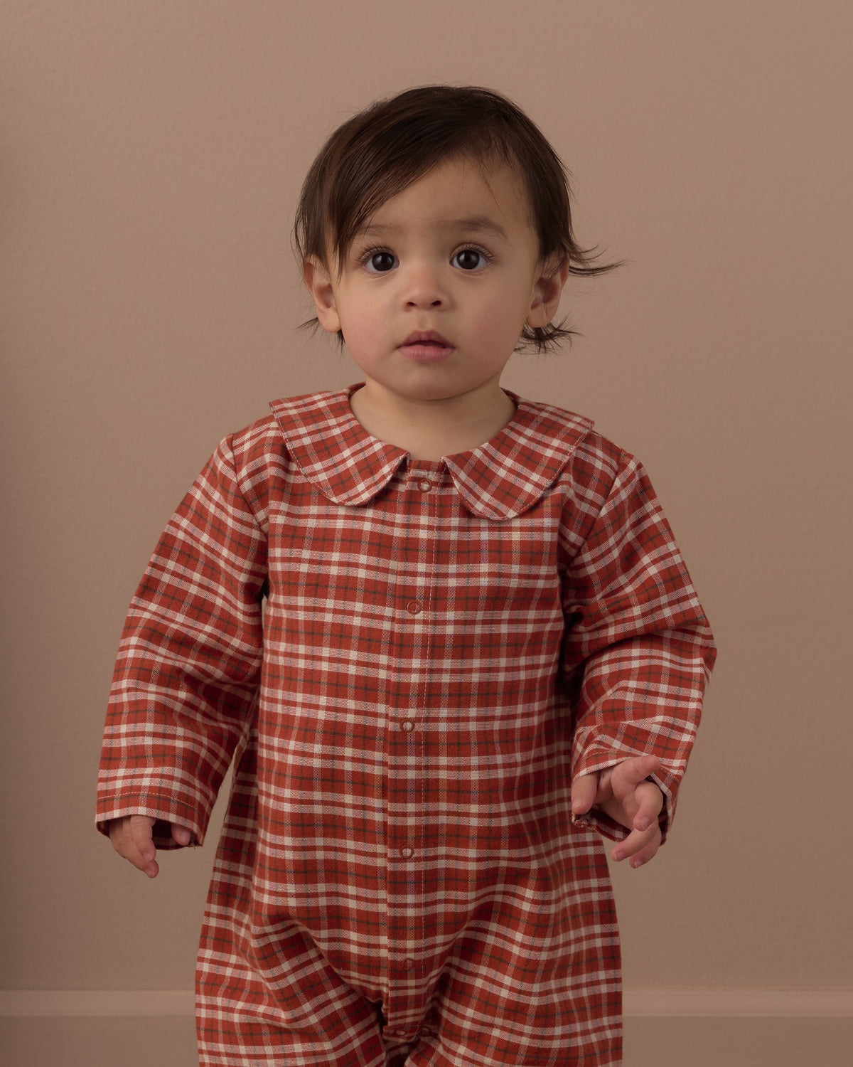 Baby wearing red checked romper with peter pan collar in front of a beige background
