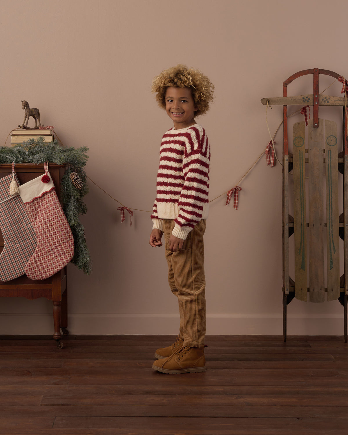 Child wearing red and white striped long sleeve sweater with brown corduroy pants and brown boots in a living room decorated for Christmas