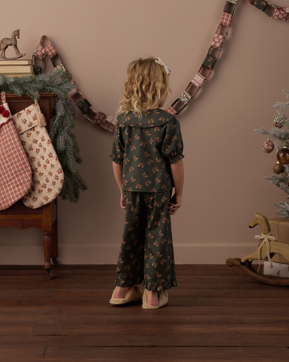 Child wearing green floral two piece pajamas in a living room decorated for Christmas, showing the back of the pajamas