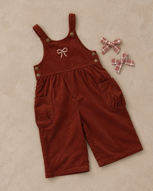 Ruby red cord overalls with a white bow embroidered on the front and side pockets, shown with two hair bows on a beige background