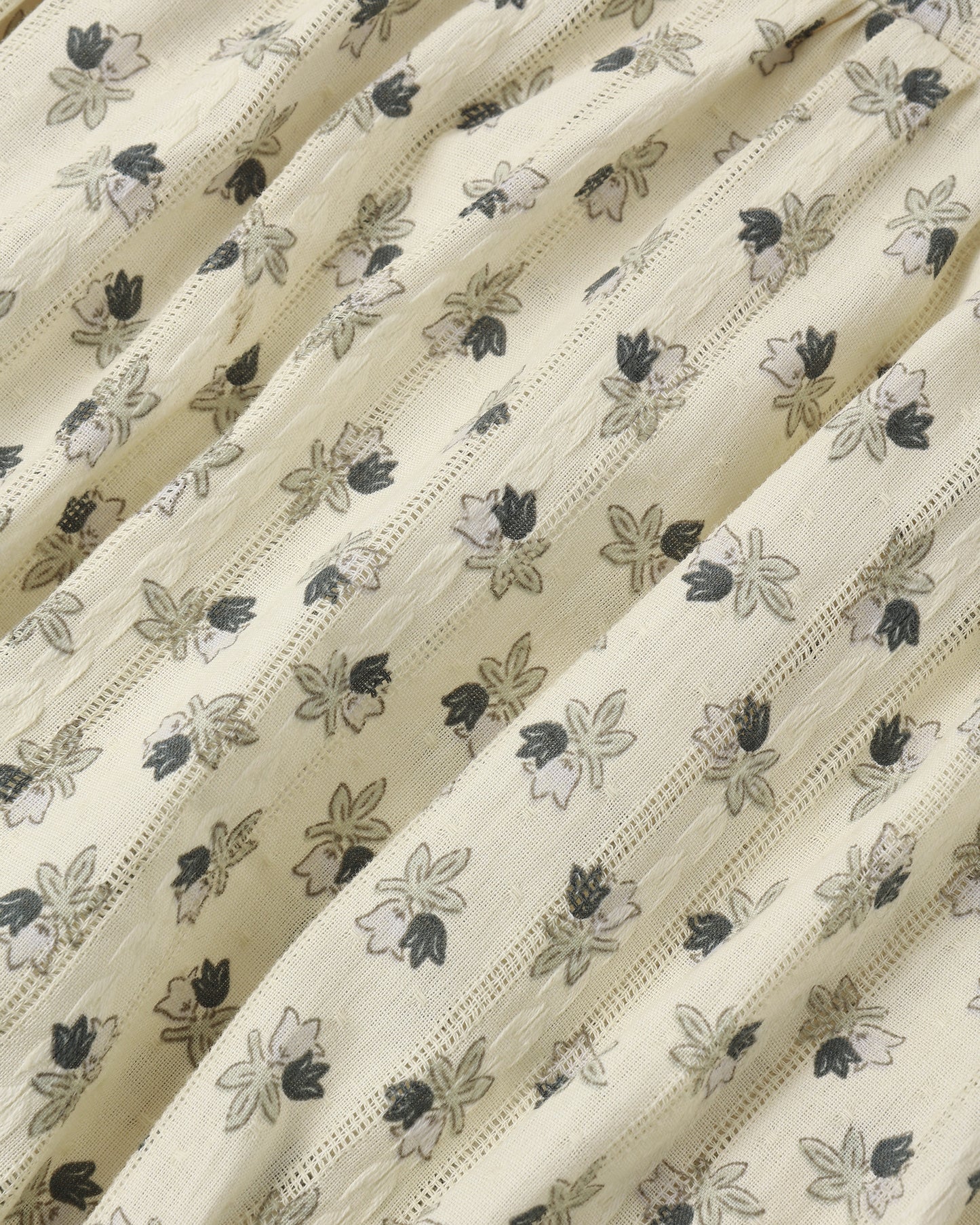 A close-up of a cream-colored fabric with a delicate, repeating floral block print in black and gray, featuring fine vertical stitched stripes.