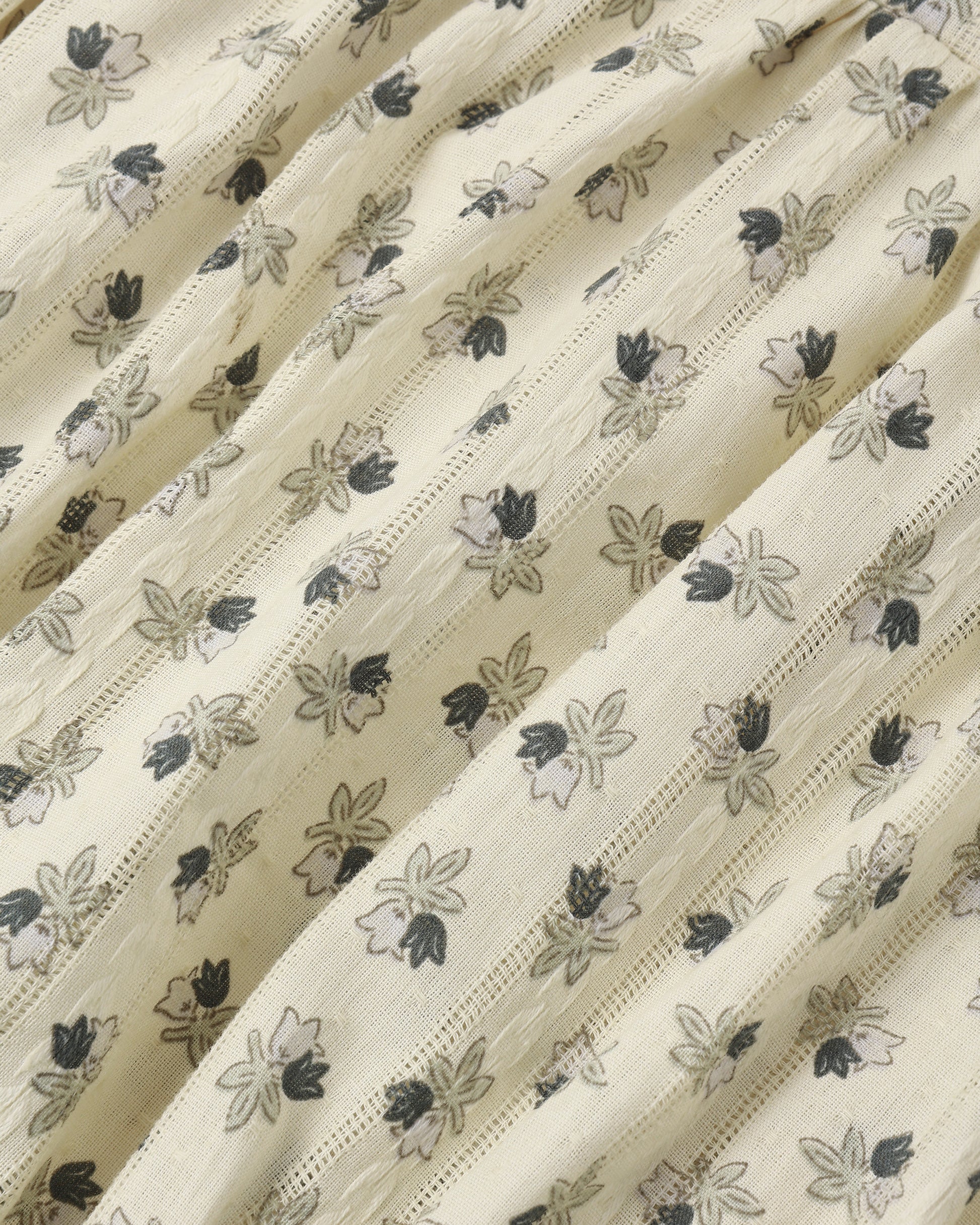 A close-up of a cream-colored fabric with a delicate, repeating floral block print in black and gray, featuring fine vertical stitched stripes.
