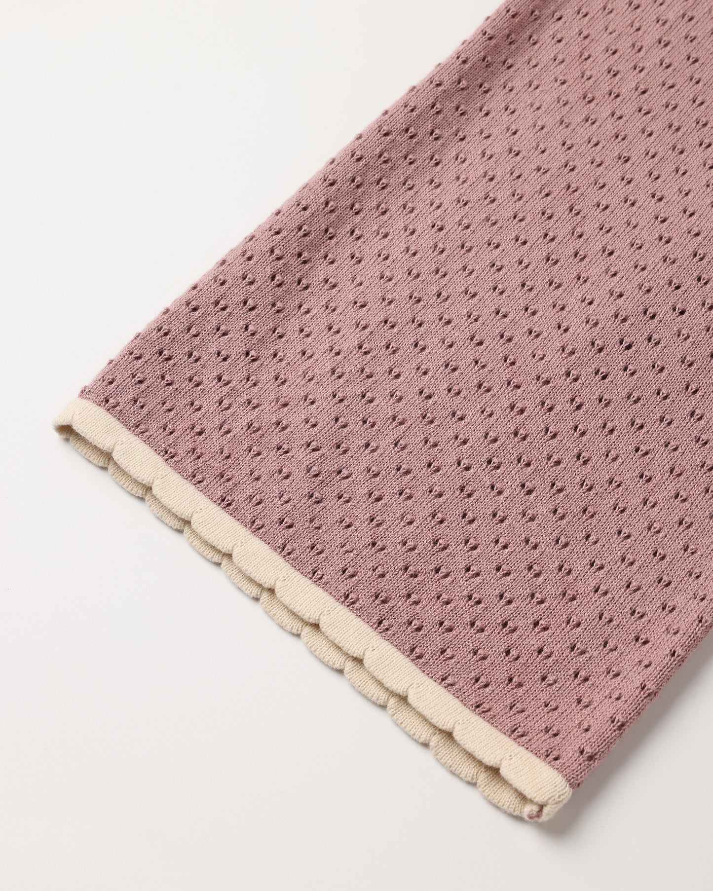 A close-up of a dusty pink textured fabric item, likely a blanket or scarf, featuring a scalloped cream border on a plain white background.
