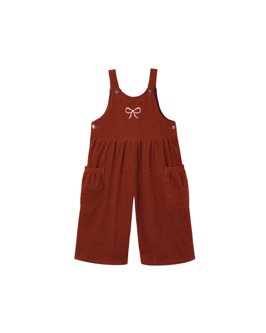 A rust-colored corduroy short overall romper with adjustable shoulder straps, side pockets, and a small white bow embroidered on the chest, shown against a plain white background.