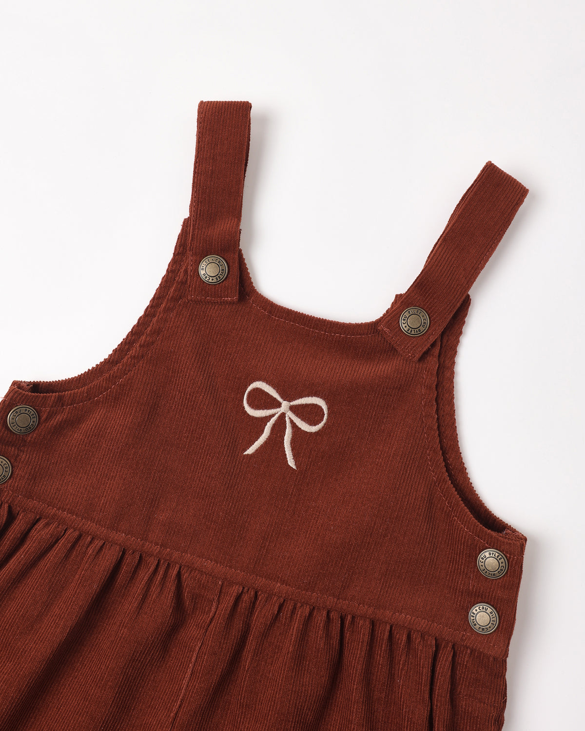 A close-up of a rust-brown corduroy baby dress or overall-style garment with wide shoulder straps, metal snap buttons, and a small white embroidered bow at the center chest, shown on a plain light background.