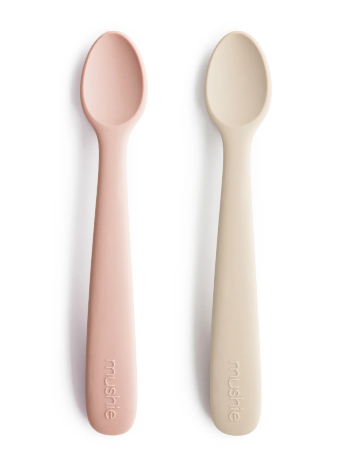 Two silicone toddler spoons, one pink and one beige, on a white background