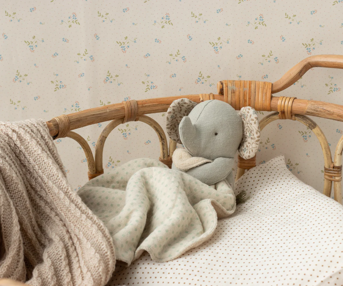 Gray elephant plush toy in a wicker crib with floral wallpaper