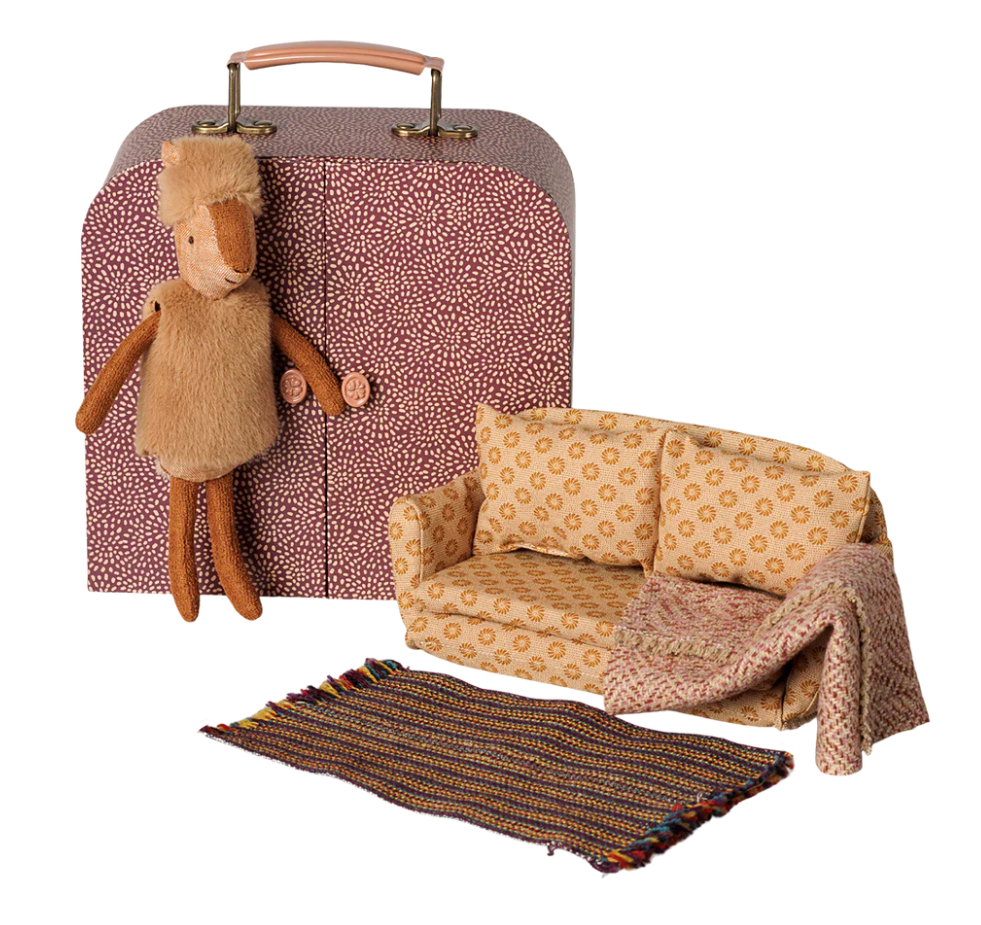 A suitcase with doors open to little sister deer dressed in a furry west and hat, a sofabed with a warm blanket and a rug
