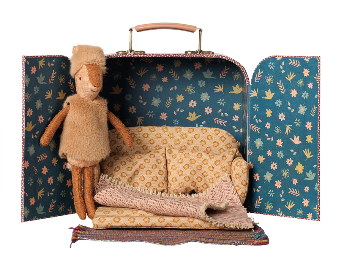 A suitcase with doors open to little sister deer dressed in a furry west and hat, a sofabed with a warm blanket and a rug
