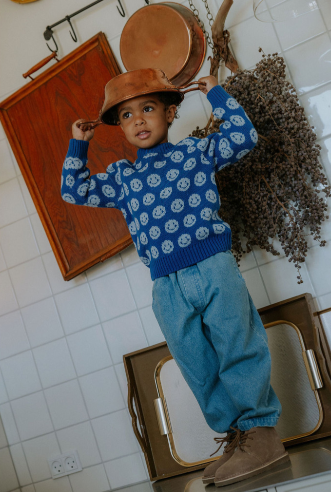 Child standing on a counter with a pan on his head, wearing denim pants and a blue smiley face sweater