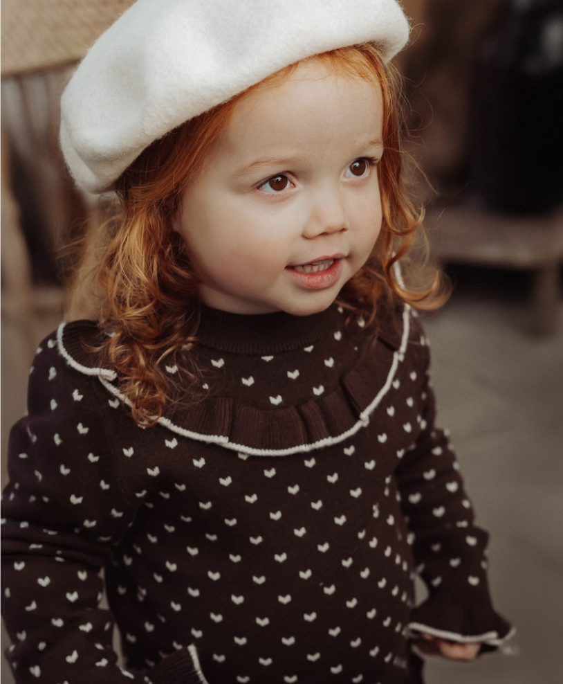 A young child wearing a dark brown knit sweater with small white motifs and a white ruffled collar trim, paired with a white beret, photographed in a softly blurred indoor setting.