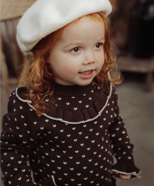 A young child wearing a dark brown knit sweater with small white motifs and a white ruffled collar trim, paired with a white beret, photographed in a softly blurred indoor setting.