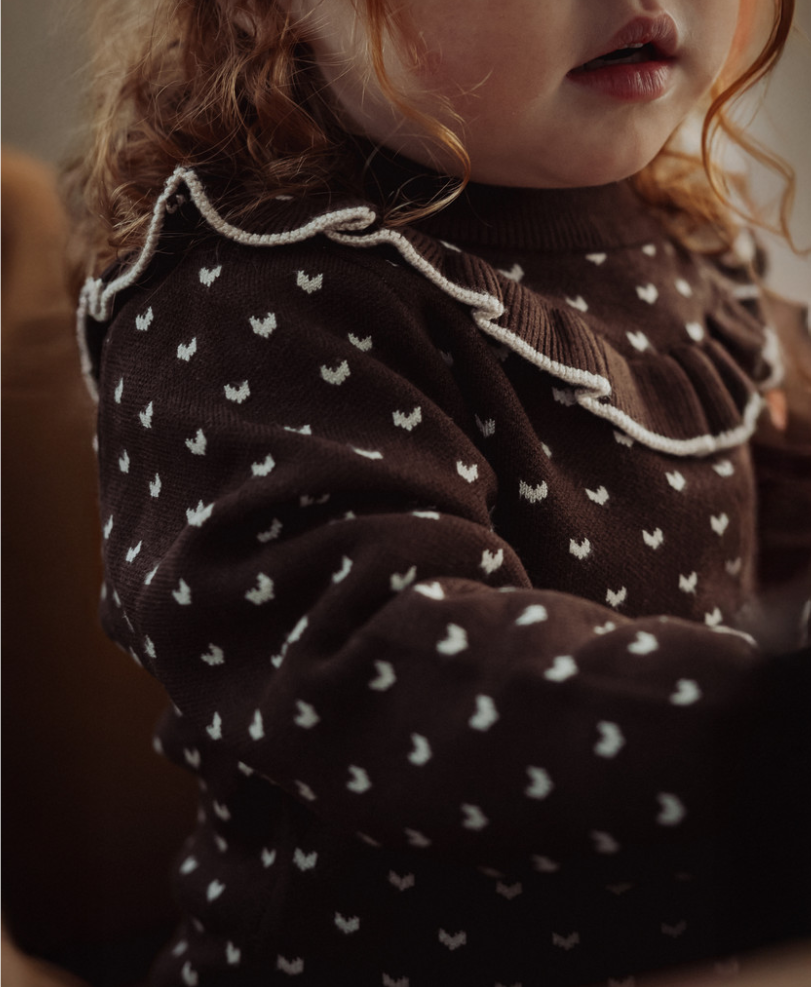 A close-up lifestyle shot of a toddler wearing a dark brown knit sweater with a white heart pattern and a white rope ruffle collar.