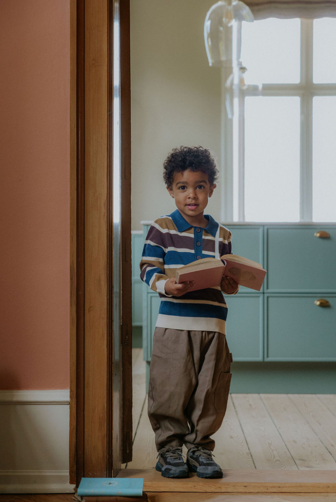 Child wearing the duna cargo pants and a striped sweater holding an antique book standing a room with a wooden floor and blue cabinet