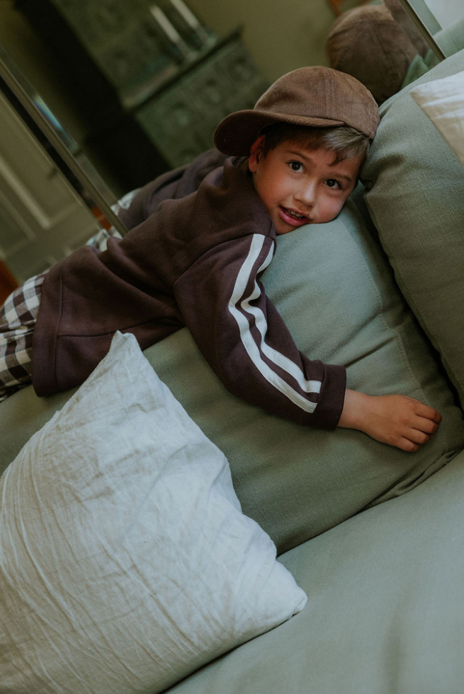 Child wearing brown cap and brown sweatshirt while laying on a couch with large pillows