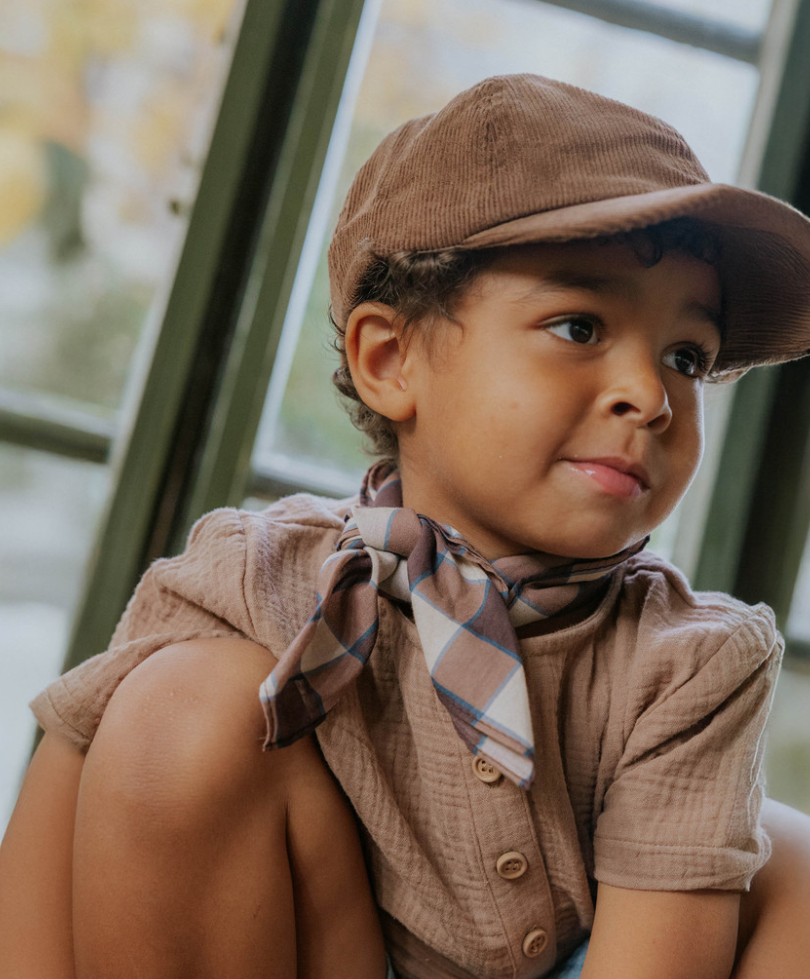 Child wearing brown cap, brown shirt and neck scarf in an interior setting