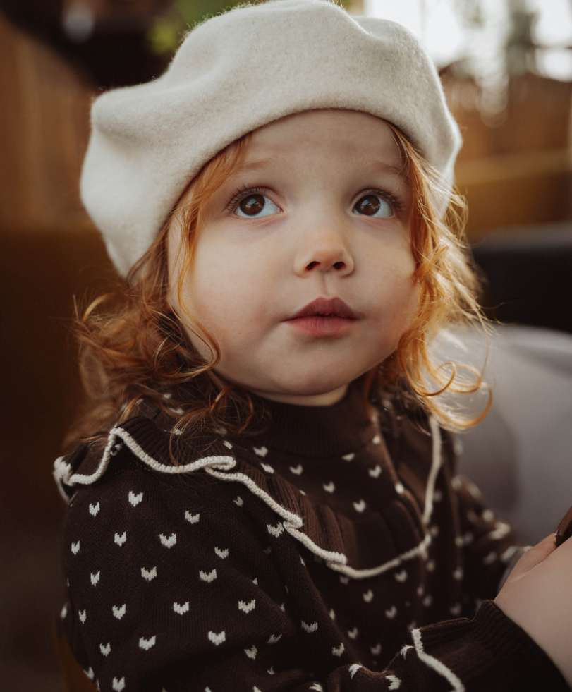Child wearing ivory beret and a brown sweater in an indoor setting