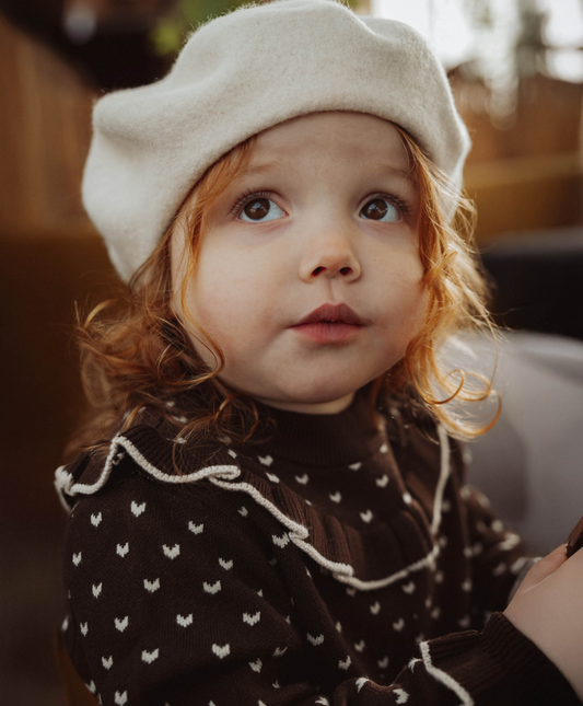 Child wearing ivory beret and a brown sweater in an indoor setting