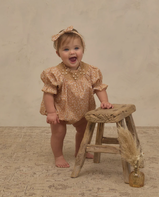 A baby girl wearing a tan and cream floral romper with a matching bow headband stands on a textured rug next to a rustic wooden stool.