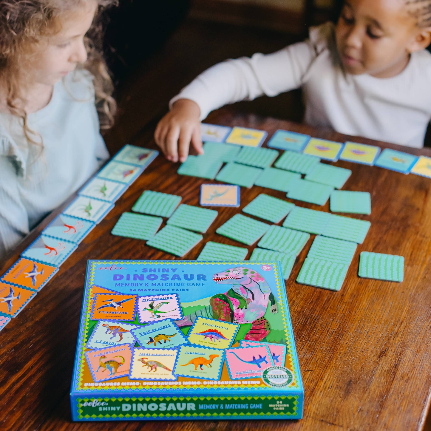 Two children playing with a Shiny Dinosaur memory and matching game on a wooden table.