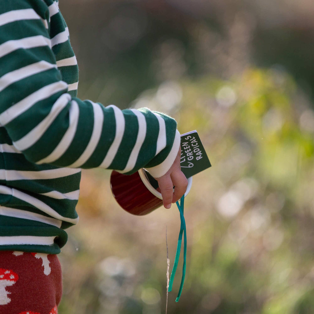 Child wearing a green and cream striped T-shirt while holding a red mug outdoors.