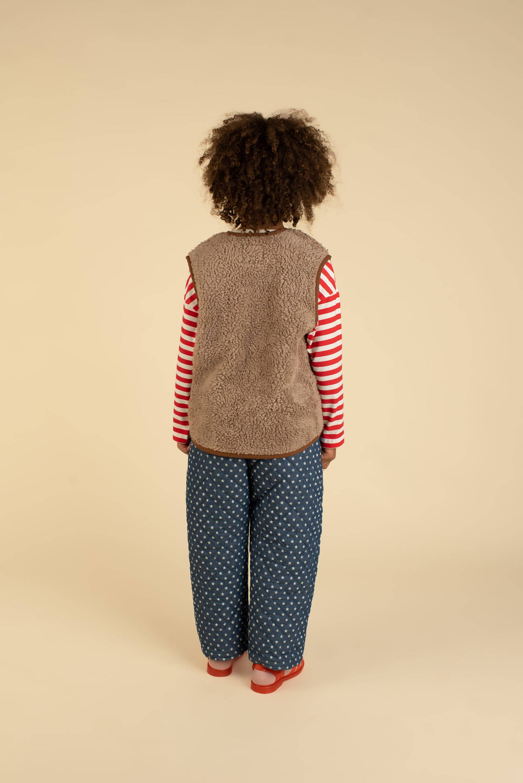 Child wearing a brown vest over a red and white striped shirt with blue patterned pants on a beige background