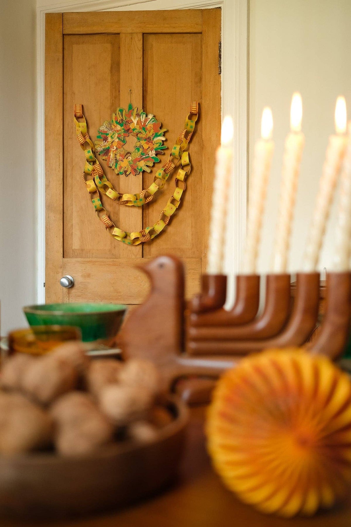 Yellow and green paper chains draped around a wreath on a wooden door, with candles in the foreground.