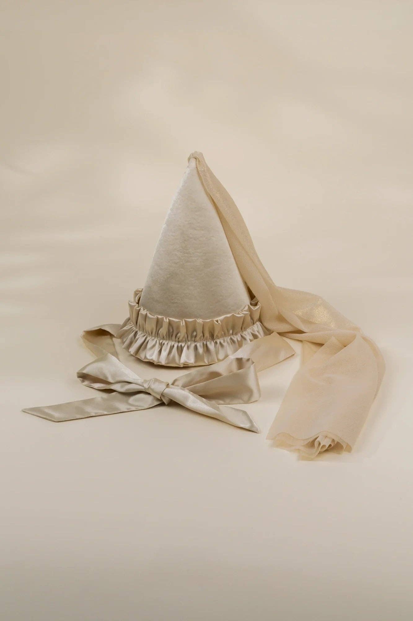 A cream-colored fabric pointed hat with a ruffled trim and long matching ribbon tails, photographed on a plain warm beige studio background.