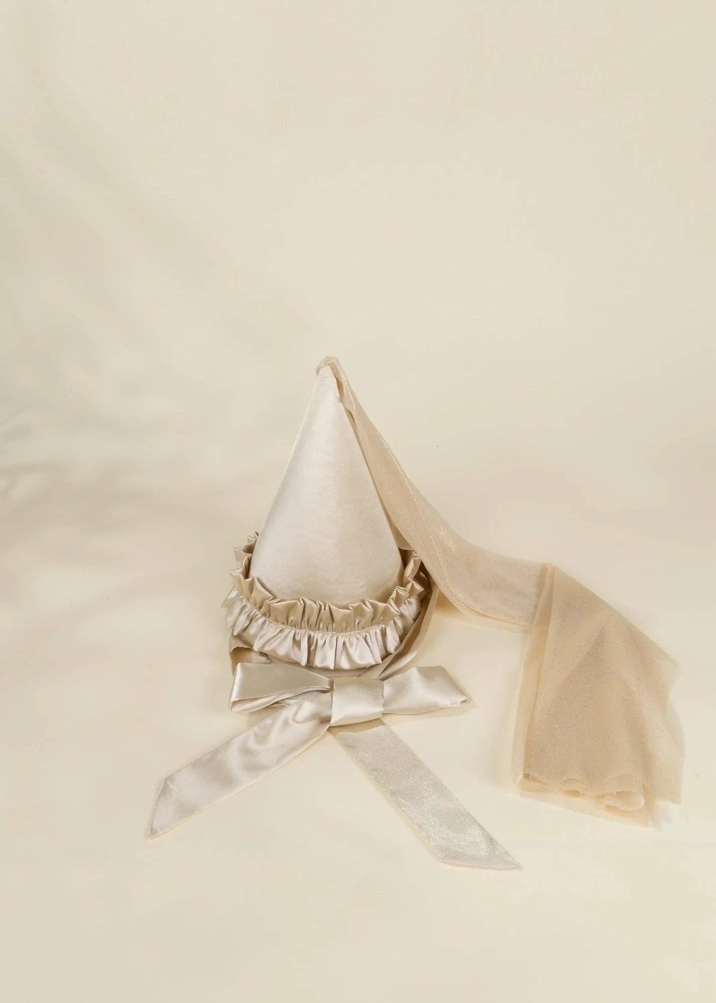 A cream-colored satin fairy hat with a ruffled trim, a matching satin bow, and a sheer tulle veil, displayed on a soft cream fabric background.