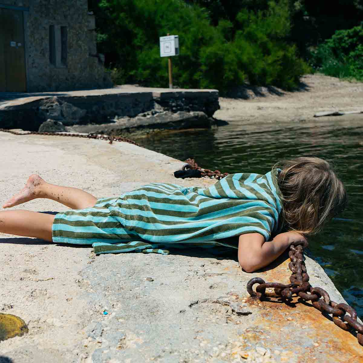Child in a striped hooded towel lying on a dock by water