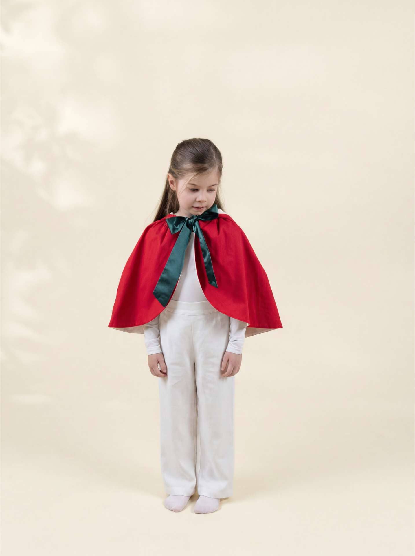 A young girl stands against a warm cream studio background wearing a bright red cape with a dark green satin neck bow over a white outfit.