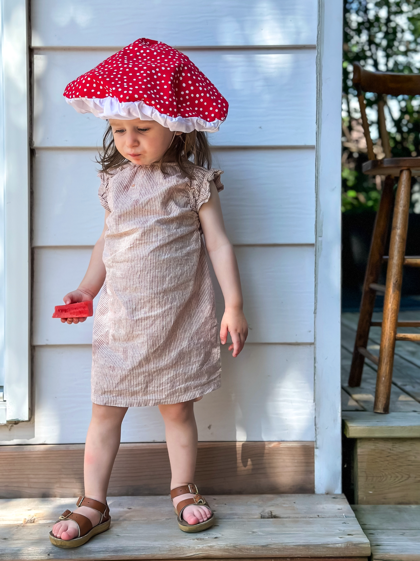 Child wearing a red polka dot mushroom hat on a wooden porch.
